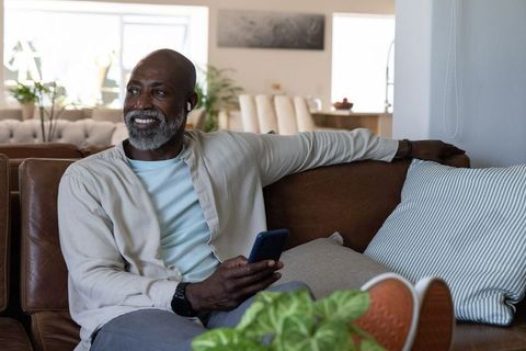 Senior african man relaxing on couch using smartphone and wireless earbuds at home