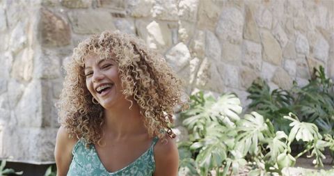 Joyful woman with curly hair laughing outdoors by stone wall