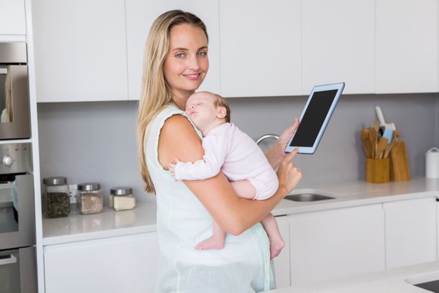 Mother holding a sleeping baby while using a digital tablet in a modern kitchen. Ideal for use in articles or advertisements about parenting, modern family life, multitasking mothers, and the integration of technology in daily routines.