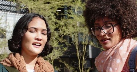 Multicultural women chatting on sunlit urban campus wearing scarves, glasses and backpack