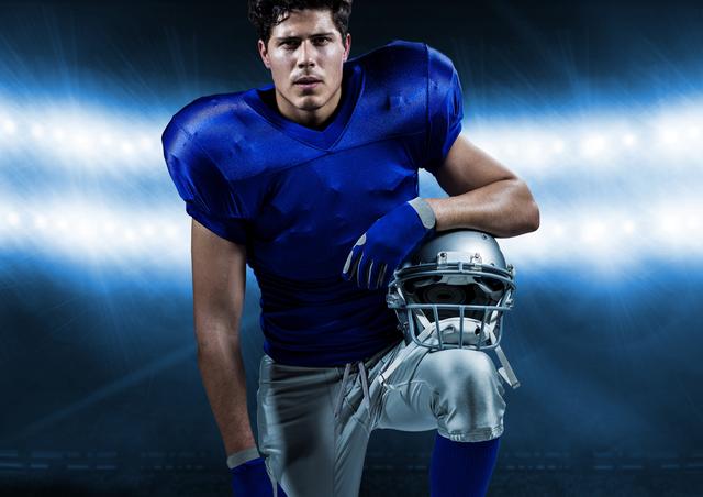 American football player wearing blue uniform and holding helmet, kneeling in a brightly lit stadium. Ideal for use in sports promotions, athletic event advertisements, and motivational posters.