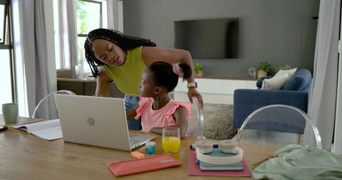 African american mother helping daughter studying on laptop at home dining table