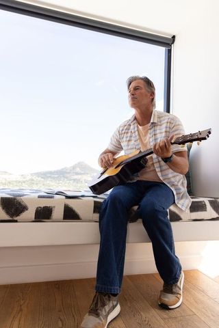 Mature man playing acoustic guitar on window bench with mountain view and sunlight