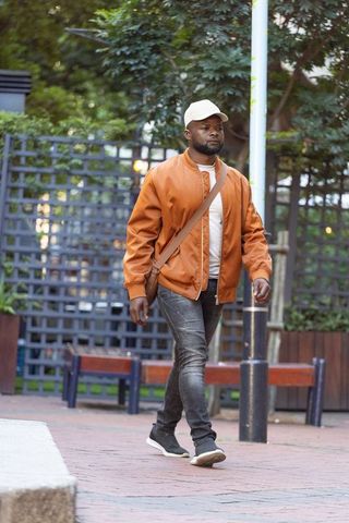 Urban man walking in brown bomber jacket and beige cap with tan crossbody bag, streetwear