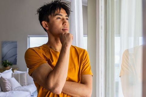 Young asian man standing by window gazing thoughtful wearing mustard tee in sunlit apartment