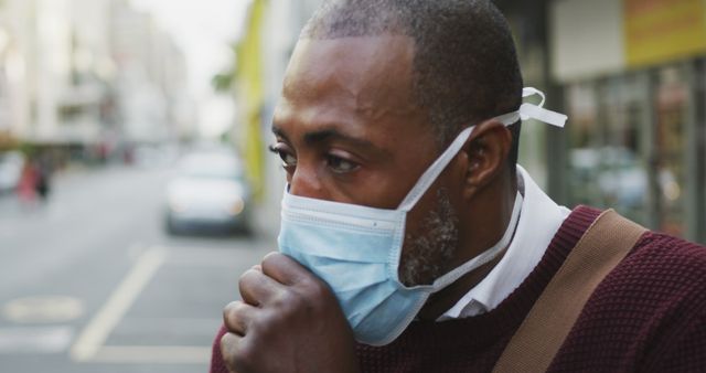 African American man out and about in the city streets during the day, wearing a face mask against air pollution and covid19 coronavirus, covering his mouth while coughing. 