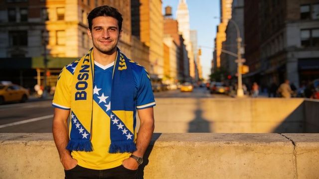 Leaning fan wearing bosnia scarf and blue jersey on sunlit urban avenue during golden hour