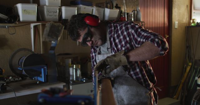 Craftsman working in a workshop with focused expression, using tools for precision woodworking. He is wearing safety goggles, ear muffs, and gloves for protection. Ideal for illustrating themes such as skilled manual labor, craftsmanship, and the importance of safety in workplaces.