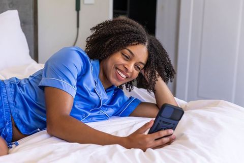 Smiling woman relaxing in blue satin pajamas on bed, texting on smartphone, cozy morning routine