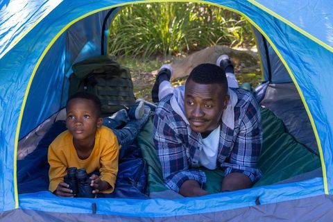 African american father camping with son in blue tent holding binoculars outdoors