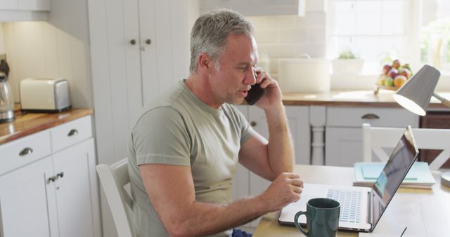 Older man multitasking by speaking on a phone call and using a laptop in a bright, modern kitchen. Ideal for depicting telecommuting, remote work, freelance profession, home office setup, lifestyle of senior citizens, and technology use in daily life.