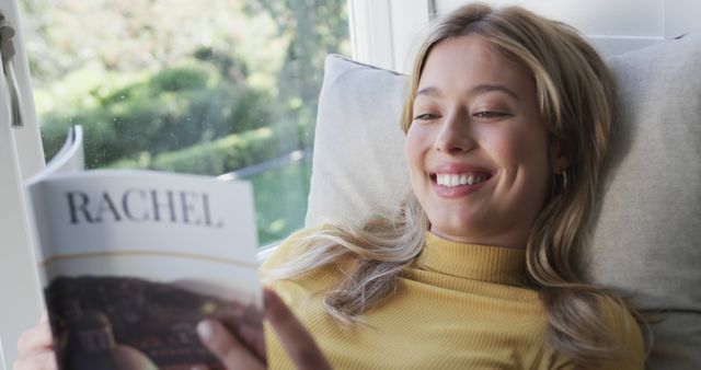 A woman relaxes while reading a book by the windowsill, with natural light pouring in. She has blond hair and is smiling, wearing a mustard-colored turtleneck. This cozy scene is ideal for promoting leisure activities, home life, or reading materials. Great for use in lifestyle displays and educational materials emphasizing relaxation and personal enjoyment.