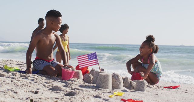 Smiling african american family building sandcastle with american flag on sunny beach. healthy, active family beach holiday.
