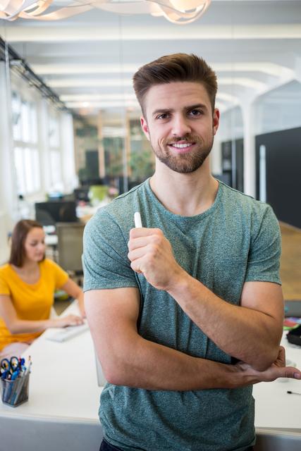 Young male business executive holding pen and smiling confidently in a modern office. Ideal for use in business, leadership, and teamwork-related content. Perfect for illustrating modern work environments, professional confidence, and collaborative spaces.