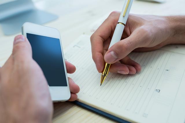 Male graphic designer writing in a diary while holding a mobile phone in an office. Ideal for use in articles or advertisements about productivity, organization, business planning, and technology in the workplace. Can also be used for illustrating concepts related to scheduling, time management, and professional work environments.
