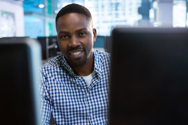 This image shows a smiling computer engineer sitting at his desk in a modern office environment. He is wearing a checkered shirt and appears to be engaged in his work. This image can be used for promoting technology services, corporate websites, IT recruitment, and business-related content.
