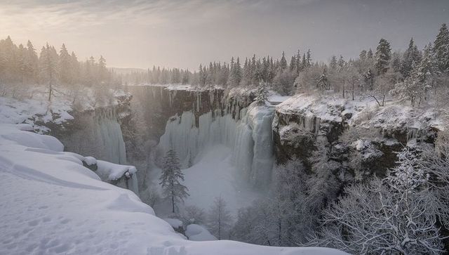 Frozen waterfall in winter canyon with snowdrifts and footprints on cliff edge at sunrise