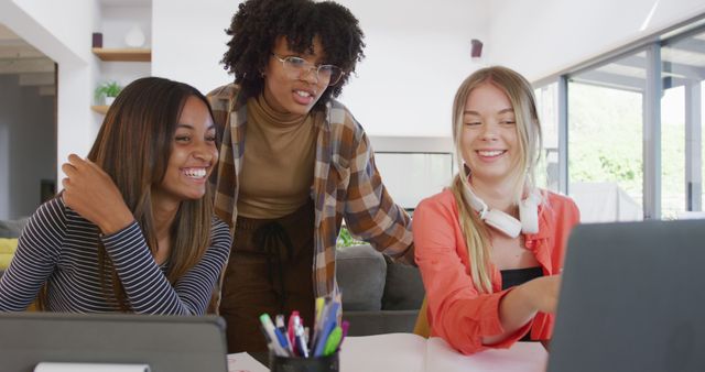 Three young women, from different ethnic backgrounds, are gathered around a table in a modern household, engaging in a collaborative task on a laptop while laughing and enjoying each other's company. This image can be used for promoting themes of diversity, teamwork, friendship, home office settings, or casual work environments.