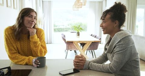 Two diverse women chatting over coffee at sunlit modern kitchen island, casual friendship