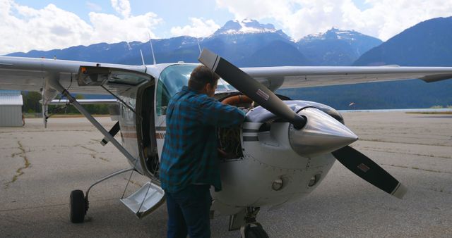 Man in blue plaid shirt examining engine of a small propeller aircraft parked on tarmac with mountains in background. Suitable for topics on aviation maintenance, aircraft technology, pilot training, travel, and outdoor adventure.