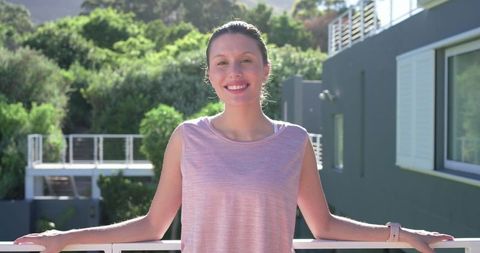 Young woman smiling on balcony in light pink top wearing smartwatch, overlooking lush hillside and d