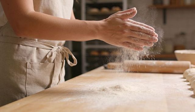 Baker dusting flour on wooden counter with rolling pin and dough rounds in linen apron