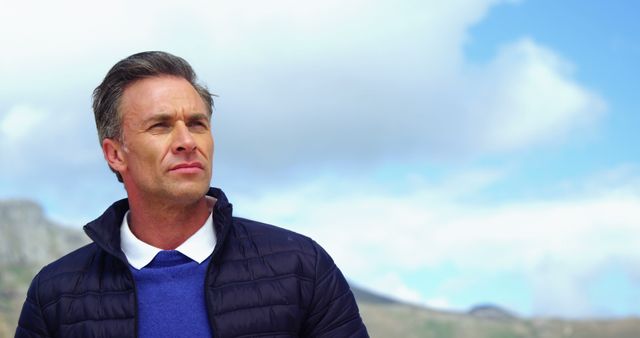 Middle-aged man looking confidently at distance, wearing casual jacket outdoors with blue sky background. Ideal for promotional materials highlighting outdoor activities, men's fashion, lifestyle concepts, personal development.