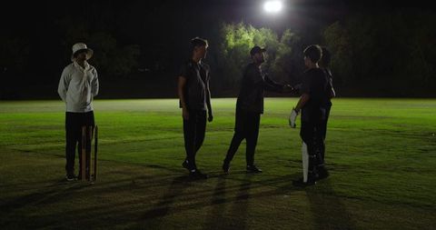 Nighttime cricket team shaking hands under floodlights on grassy pitch after training