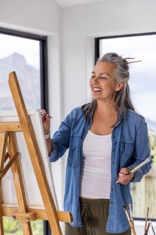 Senior woman painting on canvas in bright studio with mountain view, smiling mature artist