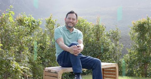 Smiling mature man sitting on wooden bench in sunny garden with trimmed hedges wearing green polo