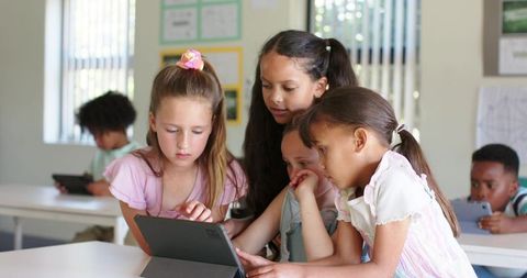 Multiracial elementary girls collaborating over tablet in bright classroom for digital learning