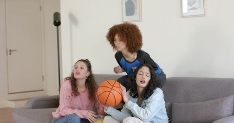 Diverse friends reacting and cheering on couch holding basketball and snacks during game