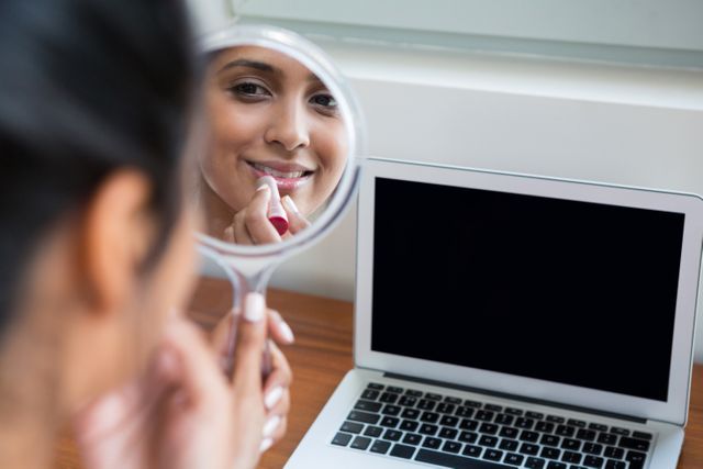 Woman applying lipstick with reflection on hand mirror by laptop at home