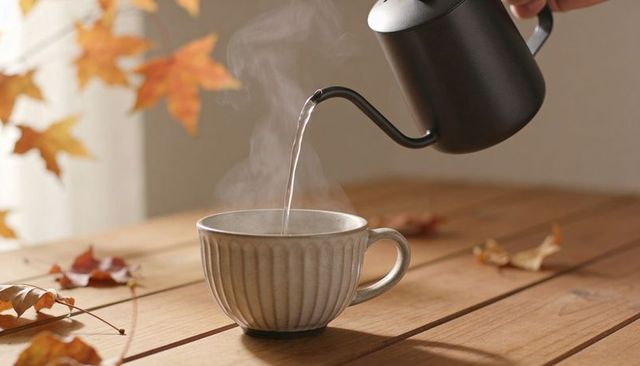 Pouring hot water from gooseneck kettle into ribbed cup on wood table with autumn leaves