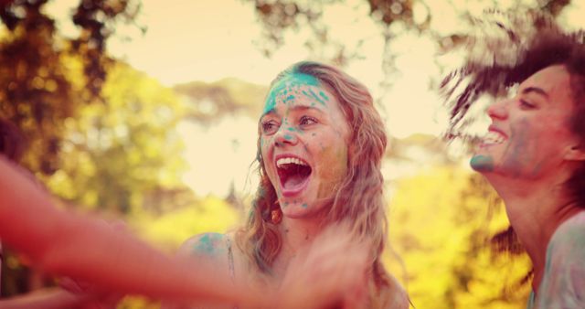 A group of young adults is engaged in a vibrant color festival, with their faces smeared with bright hues, expressing joy and excitement. Their laughter and dynamic gestures convey a sense of freedom and celebration in an outdoor setting.