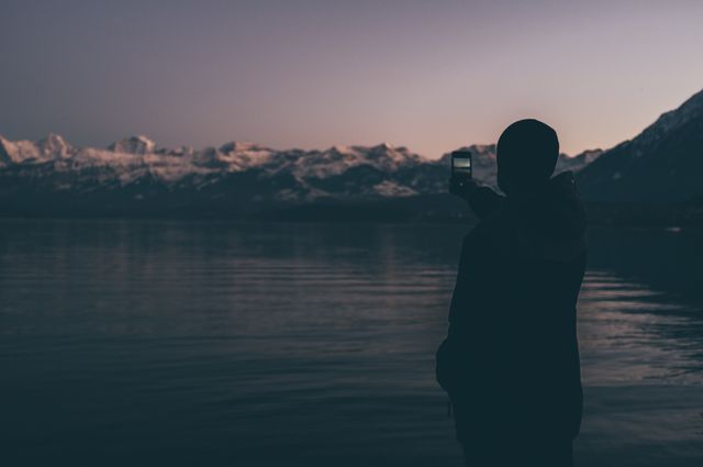 Person stands at the edge of a calm lake with mountains visible in the background, capturing the serene beauty of twilight with a smartphone. This could be used in travel blog posts, technology advertisements, motivational posters, or social media promotions related to nature photography and travel.