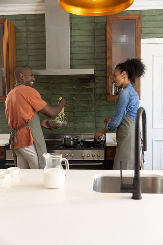 Black couple cooking pasta together in modern kitchen with green tile backsplash