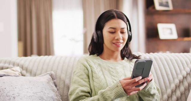 Woman sitting on sofa using smartphone while wearing headphones. Could be used for themes of technology in everyday life, leisure activity, or modern home living.