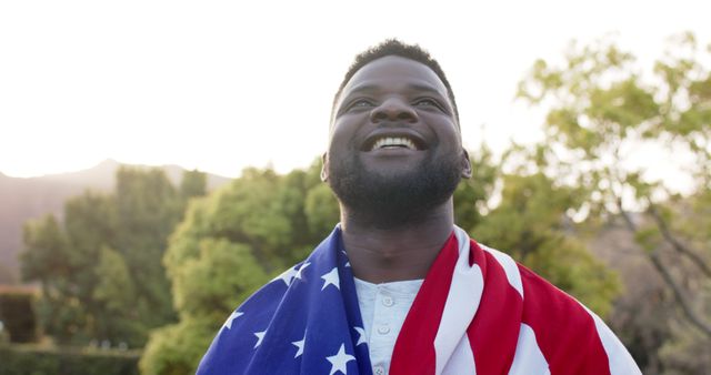 Man smiling wrapped in American flag standing outdoors. Perfect for content related to patriotism, American pride, national holidays, and cultural celebrations. Suitable for use in advertisements, social media posts, and promotional materials celebrating American identity.