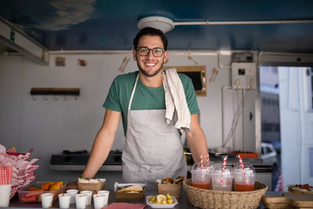 Waiter wearing apron and glasses smiling at food truck counter with various food and drinks displayed. Ideal for use in articles about street food, small businesses, customer service, and urban dining experiences.