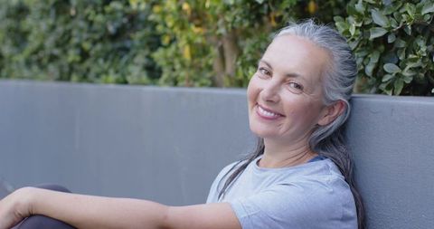 Smiling senior woman relaxing against concrete planter near trimmed hedge in courtyard
