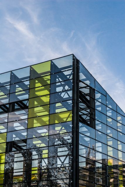 This image shows a modern glass building with reflective surfaces capturing the blue sky and light shadows around it. The light yellow details add a unique geometric pattern to the structure. This stock photo is perfect for presentations and projects related to urban development, contemporary architecture, or city planning prospects, reflecting modern design principles and minimalistic aesthetic.
