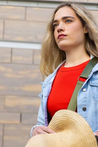 Confident young woman standing in denim jacket holding straw hat, urban fashion portrait