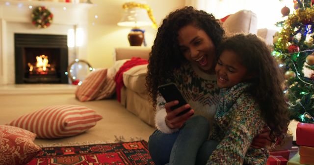 Mother and daughter sitting together on floor by fireplace, smiling and looking at smartphone. Living room decorated for holidays with Christmas tree and lights. Perfect for family, holiday, and lifestyle content, showcasing festive traditions and cozy interiors.