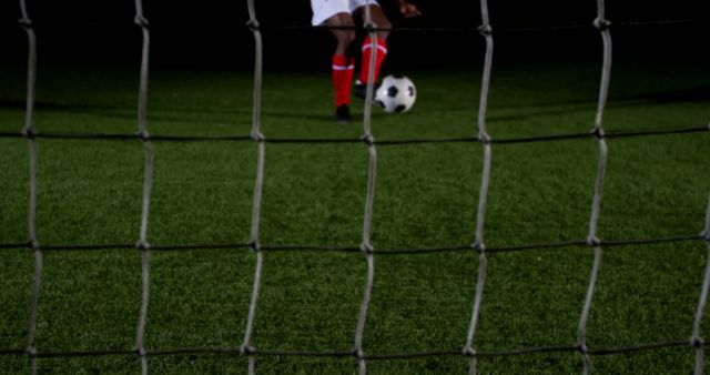 A soccer player in red and white gear is about to kick a ball on a lush green pitch, viewed from behind the goal net. Capturing the anticipation of a potential goal, the image focuses on the player's action and the iconic black and white soccer ball.