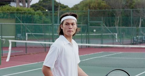 Asian tennis player standing on outdoor hard court holding racket wearing white polo and headband