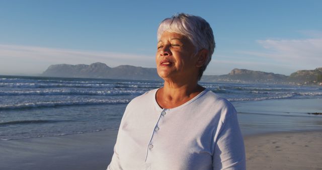 Senior woman enjoying a peaceful moment on the beach at sunrise. Ideal for use in promotions related to retirement, wellness, tranquility, relaxation, and coastal living. This image depicts calmness and the beauty of connecting with nature at an elderly age.