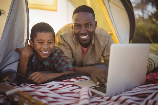 Father and son enjoying quality time together in a tent, using a laptop. Perfect for themes related to family bonding, outdoor activities, technology in nature, and happy moments. Ideal for advertisements, blog posts, and articles focusing on family life, camping trips, and modern parenting.