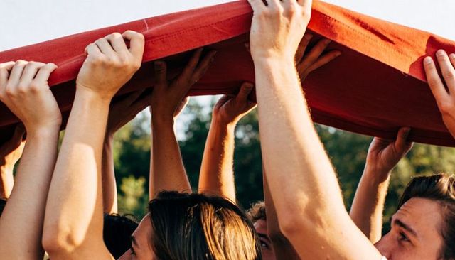 Young friends raising red fabric overhead in sunny park celebrating teamwork, unity, summer fun