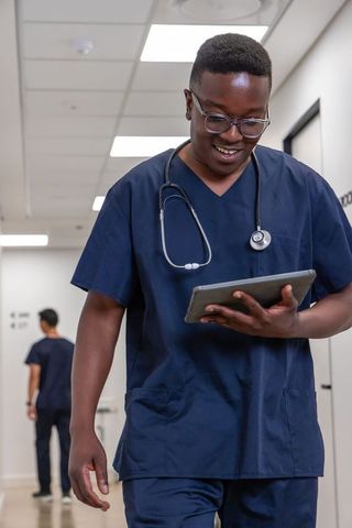 Smiling nurse walking clinic corridor using tablet with stethoscope for digital care